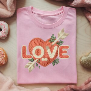 May include: A light pink t-shirt with a crew neck featuring a heart design with the word "LOVE" in a cream color. The heart is pierced by an arrow and surrounded by green leaves. The shirt is a Valentine's Day theme.