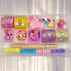 May include: A clear plastic organizer filled with colorful play dough, candies, and accessories. The play dough is decorated with sprinkles and candies. A wooden rolling pin and colorful pipe cleaners are also included.