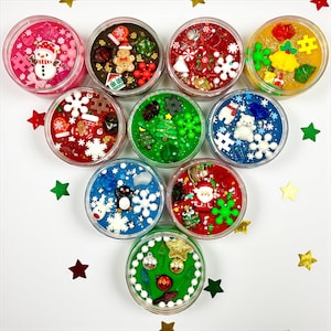 May include: A collection of clear plastic containers filled with colorful slime, each decorated with festive Christmas-themed embellishments. Decorations include snowmen, gingerbread men, snowflakes, and miniature Christmas trees. The containers are arranged on a white surface with scattered star-shaped confetti.
