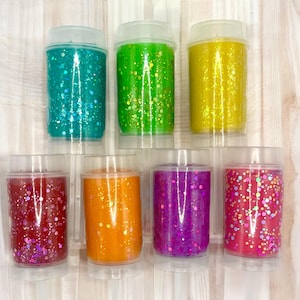 May include: Seven clear plastic containers filled with colorful glitter in shades of teal, green, yellow, red, orange, purple, and pink. Each container has a clear lid and is displayed on a white surface.