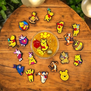 May include: A collection of Winnie the Pooh character charms, featuring various poses and designs, including a honey pot charm with the word "Honey" on it. The charms are displayed on a wooden surface with a bowl of yellow slime in the center.
