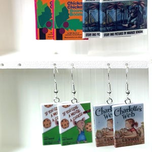 Op de afbeelding: Vier paar oorbellen in de vorm van miniatuurboeken met klassieke kinderboekcovers zoals Chicka Chicka Boom Boom, Where the Wild Things Are, If You Give a Mouse a Cookie en Charlotte's Web, bevestigd aan zilverkleurige metalen oorhaken.