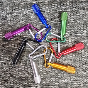 Puede incluir: Surtido de pequeñas linternas de colores con mosquetones. Las linternas son de varios colores, incluyendo azul, morado, negro, plateado, verde, rojo y dorado. Las linternas tienen un agarre texturizado y están unidas a los mosquetones.