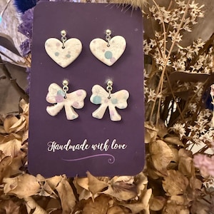 May include: A pair of heart and bow earrings displayed on a purple card. The earrings are white with colourful speckles. The card reads "Handmade with love" in cursive.