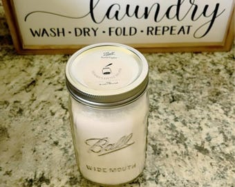 Puede incluir: Un tarro de vidrio transparente con una tapa blanca y una etiqueta blanca que dice "Ball Wide Mouth". La etiqueta también tiene una pequeña imagen de una cuchara y el texto "Laundry Little Helper". El tarro está lleno de un polvo blanco y está sentado en una encimera marrón claro.