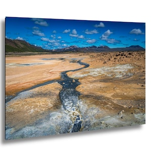 May include: A landscape photograph showing a dark, winding stream cutting through a vast, arid terrain of tan and grey earth. In the distance, dark mountain ranges sit under a bright blue sky filled with scattered white clouds.