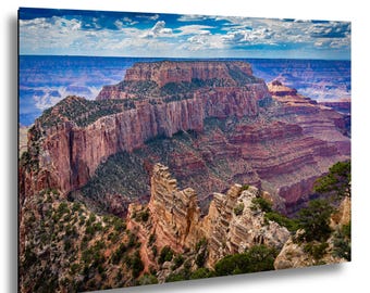May include: A landscape photograph of the Grand Canyon, showcasing layered rock formations in shades of red, brown, and tan. The sky is a vibrant blue with scattered white clouds. Green vegetation dots the canyon's edges.