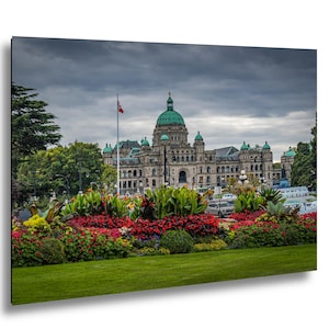 Puede incluir: Una fotografía de los edificios del Parlamento de la Columbia Británica en Victoria, Canadá. El edificio tiene una gran cúpula y varias cúpulas más pequeñas, todas con tejados verdes. Un colorido parterre de flores está en primer plano y un cielo nublado encima.