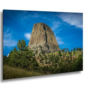 May include: A landscape photograph of Devils Tower National Monument in Wyoming. The image shows the tower's distinctive rock formation against a bright blue sky with scattered clouds. Green trees and vegetation are in the foreground.