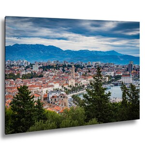 Może przedstawiać: Krajobrazowy wydruk nadmorskiego miasta z czerwonymi dachówkami, katedrą i portem. Góry w tle pod zachmurzonym niebem. Zielone drzewa kadrują pierwszy plan.