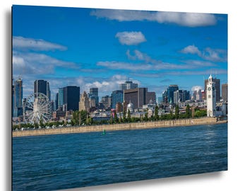 Puede incluir: Una vista panorámica de un horizonte urbano con una noria, una torre del reloj y varios edificios bajo un cielo azul brillante con nubes blancas. El primer plano presenta una masa de agua.