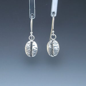 May include: A pair of silver dangle earrings featuring a coffee bean design. The earrings are made from sterling silver and have a textured finish.
