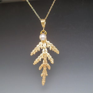 May include: A gold necklace with a delicate, detailed pendant shaped like a fern frond. A single pearl hangs from the top of the pendant.