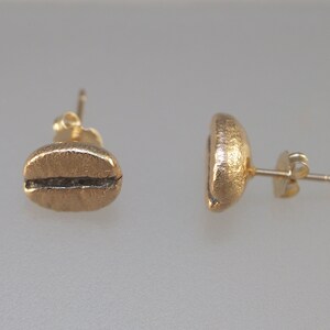 May include: A pair of gold-coloured stud earrings in the shape of coffee beans. Each earring has a textured surface and a dark line detail representing the bean's split. The earrings are shown against a light background.