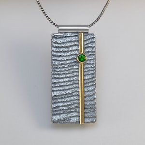 May include: A silver and gold pendant necklace with a textured rectangular pendant and a green gemstone. The pendant has a vertical gold bar in the center.