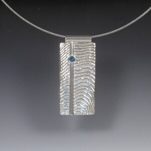 May include: A silver pendant necklace with a textured rectangular pendant featuring a blue gemstone. The pendant has a vertical line down the center.