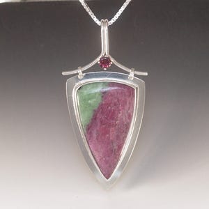 May include: A silver pendant necklace with a shield-shaped gemstone, displaying green and magenta colours. A small, round, red gemstone is set above the shield. The pendant hangs from a delicate silver chain.