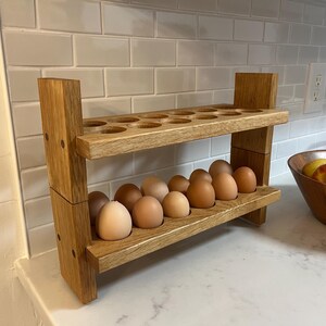 May include: A two-tiered wooden egg holder with a natural finish. The top tier has circular cutouts for eggs, while the bottom tier holds a dozen brown eggs. The holder is on a white countertop, with a bowl of fruit in the background.