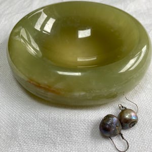 May include: A smooth, round, light green stone bowl with a polished finish and a subtle orange vein, accompanied by a pair of dark, iridescent, round drop earrings with silver-toned metal hooks and a textured, organic surface appearance.