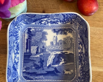 May include: A square, blue and white ceramic dish with a detailed landscape scene in the center. The rim features a floral and scroll design. Two red plums sit nearby on a wooden surface.