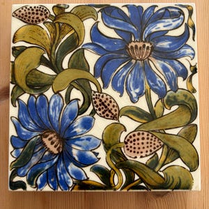 May include: A square ceramic tile featuring a hand-painted floral pattern with two large blue flowers, winding green leaves, and small spotted seed pods on a cream-coloured background with a visible crackle glaze finish throughout the surface.