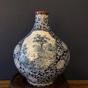 May include: A blue and white ceramic vase with a landscape scene and geometric patterns. The vase has a brown rim and sits on a wooden stand. The landscape scene features trees and a small figure.