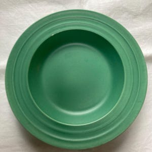 May include: A green ceramic bowl with a deep, rounded shape and a slightly raised rim. The bowl has a smooth, glossy finish and a subtle, textured design.