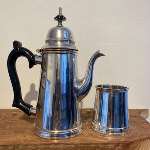May include: A silver coffee pot with a black handle and a silver creamer. The coffee pot has a domed lid with a black knob.