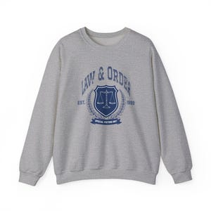 May include: A heather gray crewneck sweatshirt featuring a dark blue graphic on the chest. The design includes the text LAW & ORDER, EST. 1999, and SPECIAL VICTIMS UNIT, surrounding a shield emblem with a scale of justice and laurel leaf accents.