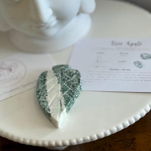 May include: A leaf-shaped stone carving in green and white, displayed on a white plate. The stone features intricate veining. A white Buddha head statue is in the background, alongside a card with text.