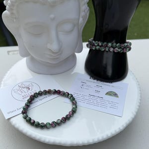 May include: A white ceramic Buddha head and two bracelets with green and pink beads. One bracelet rests on a white plate, and the other is displayed on a black hand-shaped display. A card with text is also on the plate.