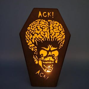 May include: A wooden coffin-shaped lamp with a cutout of an alien head and the word "ACK!" on the front. The lamp is lit from inside, casting a warm glow.