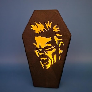 May include: A wooden coffin-shaped night light with a cutout of a man's face with spiky hair. The light is on and glowing yellow.
