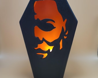 May include: A black wooden coffin-shaped candle holder with a cutout of a face. The face is illuminated from within, creating a spooky Halloween effect.