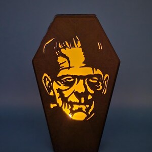 May include: A wooden coffin-shaped lamp with a cutout of Frankenstein's face. The lamp is lit from the inside, casting a warm glow.