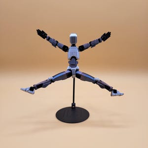 May include: A gray and black articulated action figure with a white head and black hands. The figure is posed with its legs spread wide and its arms raised above its head. The figure is standing on a black circular base.