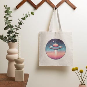 May include: A cream-colored tote bag with a circular graphic of a retro-style UFO emitting a beam of light. The design features a pink and blue color scheme with stars and a planet. The bag is hanging on a wooden wall rack.