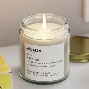 Puede incluir: Una vela encendida en un frasco de vidrio transparente con una etiqueta blanca. La etiqueta dice "MICAELA" seguida de una definición, incluyendo "Boy mom". La vela es de 113g y tiene un tiempo de combustión de 20 horas.