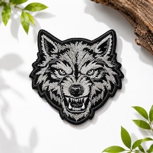 May include: An embroidered patch featuring the head of a snarling wolf. The design uses black, white, and gray thread to create a detailed, high-contrast image of the animal with bared teeth, pointed ears, and intense eyes, finished with a black border.