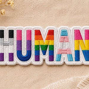 May include: Embroidered patch with the word "HUMAN" in rainbow colors, representing various LGBTQ+ pride flags. The patch has a white border and is set against a neutral fabric background.