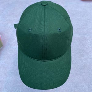 May include: A solid green baseball cap with a curved brim. The cap has a button on top and two small eyelets on the crown. The adjustable strap is visible on the back. The cap is made of a durable fabric.