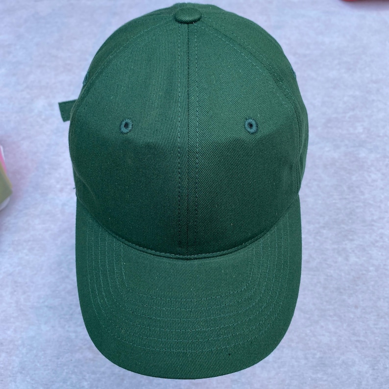 May include: A solid green baseball cap with a curved brim. The cap has a button on top and two small eyelets on the crown. The adjustable strap is visible on the back. The cap is made of a durable fabric.