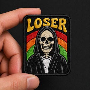 May include: Embroidered patch featuring a skull in a black hooded robe, with the word "LOSER" in yellow above a rainbow design. The patch is rectangular with a black border and is held by a hand.