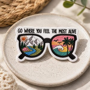 May include: An embroidered patch shaped like eyeglasses with the text GO WHERE YOU FEEL THE MOST ALIVE. The left lens shows a mountain landscape with a river and tent, while the right lens depicts a tropical beach scene with palm trees and waves.