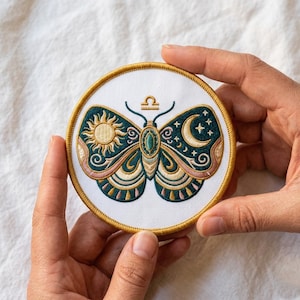 May include: Embroidered patch with a butterfly design and Libra symbol. The butterfly features a sun and moon design with teal, gold, and pink accents. The round patch has a gold border. It is held by two hands.