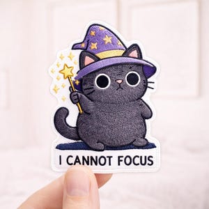 May include: Embroidered patch featuring a cartoon black cat wearing a purple wizard hat with gold stars. The cat holds a gold wand with a star-shaped tip. The text "I CANNOT FOCUS" is at the bottom of the patch.