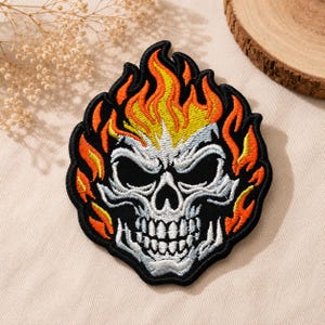 May include: An embroidered patch featuring a skull with a black base, white and light blue facial details, and vibrant yellow and orange flames rising from the top and sides. The design is outlined in black thread with a solid black border.