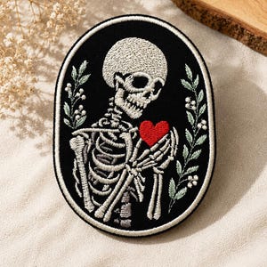 May include: An oval-shaped embroidered patch featuring a white skeleton on a black background. The skeleton holds a small, bright red heart in its hand. The design is framed by a white border and flanked by two delicate, light green leafy branches.