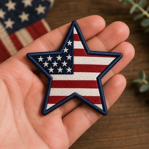 May include: Embroidered star patch featuring the American flag design. The patch has a blue border, white stars on a blue background, and red and white stripes. The patch is held in a hand.