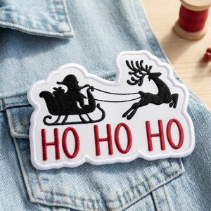 May include: A white embroidered patch featuring a black silhouette of Santa Claus in a sleigh pulled by a reindeer. The text HO HO HO is embroidered in red below the figures. The patch has a scalloped white border and is placed on denim fabric.